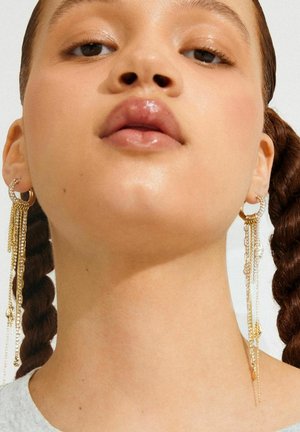 Primer plano de una mujer con el cabello trenzado que lleva pendientes largos de cadena dorada y labios brillantes, mirando ligeramente hacia arriba contra un fondo liso.