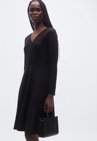 Must pikkade varrukatega kleit V-kaelusega, keha järgi istuv ülakeha ja lehvikseelikuosaga. Modell hoiab väikest musta tekstuuriga käekotti. Õrnalt mustriline sile kangas.