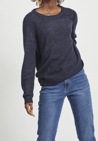 Femme portant un pull bleu marine sombre et un jean bleu, affichant une tenue décontractée avec de grandes boucles d'oreilles en bois rondes visibles.