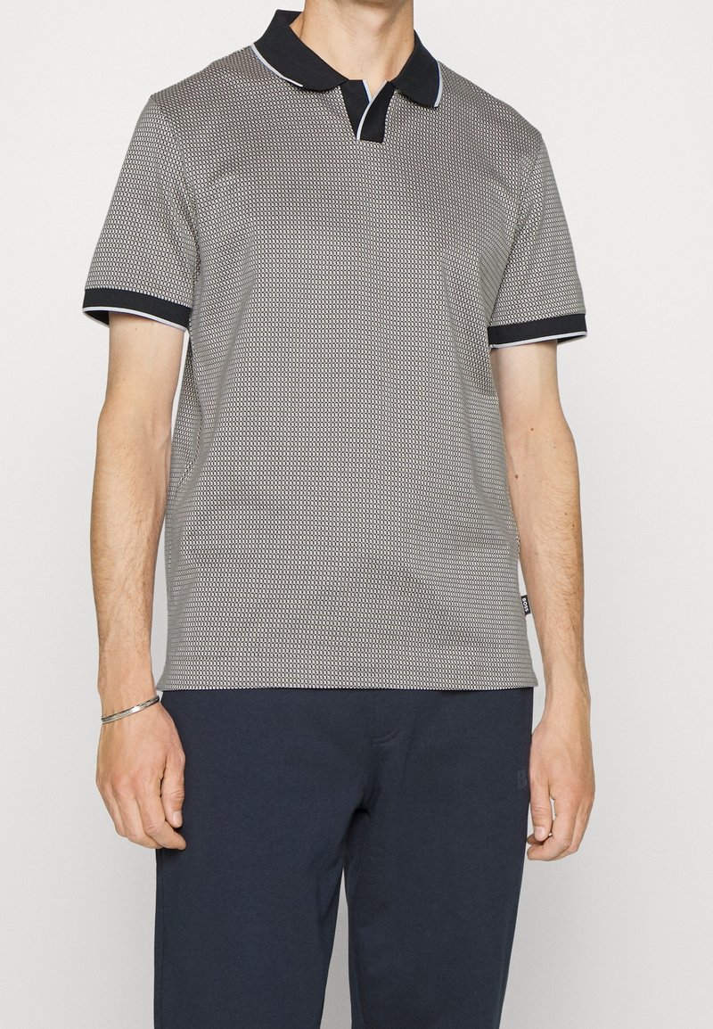 Homme portant un polo gris à manches courtes avec motif, col et poignets noirs, et un pantalon foncé sur un fond uni.