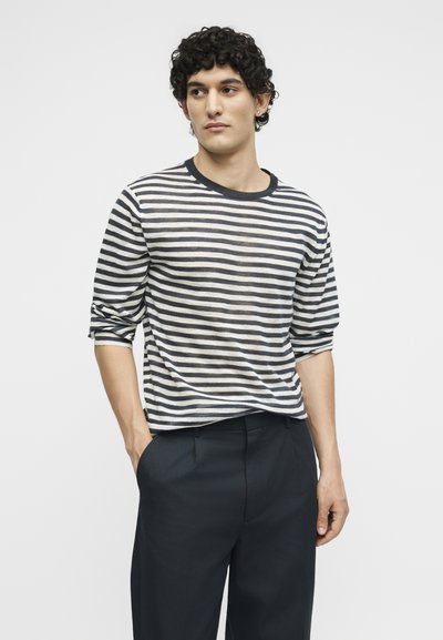 Νέος άνδρας με σγουρά μαλλιά που φοράει μακρυμάνικη μαύρο-άσπρη ριγέ μπλούζα και σκούρο παντελόνι, στέκεται με το ένα χέρι στην τσέπη.