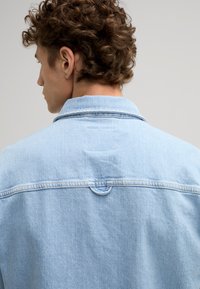 Giacca in denim azzurro chiaro con colletto classico, dettagli cucitura sul retro e un piccolo anello decorativo nella zona del collo.