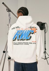 Fehér kapucnis pulóver, amelyen egy nagy kék "RWC" grafika található narancssárga kiemelésekkel és felirattal. Az anyag lágy tapintású. Sötét farmernadrággal viselve.