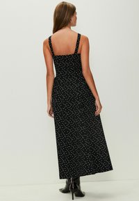 Black polka dot maxi dress with wide straps, fitted bodice, and a flared skirt. Smooth texture; worn with black heeled boots.