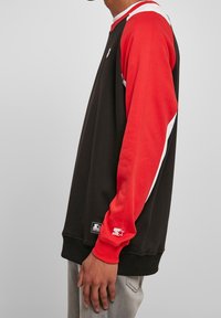 Persona vestida con un sudadera negra de mangas rojas y blancas y pantalones grises, de pie de lado contra un fondo liso.