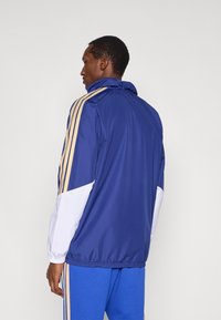 Tmavomodrá vetrovka s bielymi a zlatými akcentmi, vybavená vysokým golierom, elastickými manžetami a voľnejším strihom. Výrazný trojpásový dizajn na rukávoch.