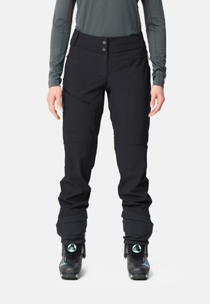Femme portant un pantalon de ski noir, des chaussures de ski et un haut gris à manches longues, debout devant un fond clair uni.