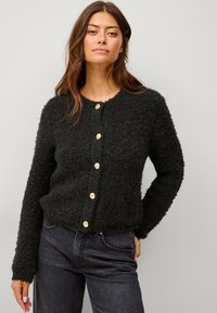 Schwarze, flauschige Strickjacke mit Rundhalsausschnitt und sechs goldfarbigen Knöpfen auf der Vorderseite. Lange Ärmel und eine weiche Textur. Getragen über blauen Jeans.