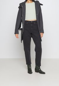 Grå ullrock med huva, ljusgrön crop top, svarta högmidjade jeans och gröna ankelboots med krokodilmönster, stående framför en neutral bakgrund.