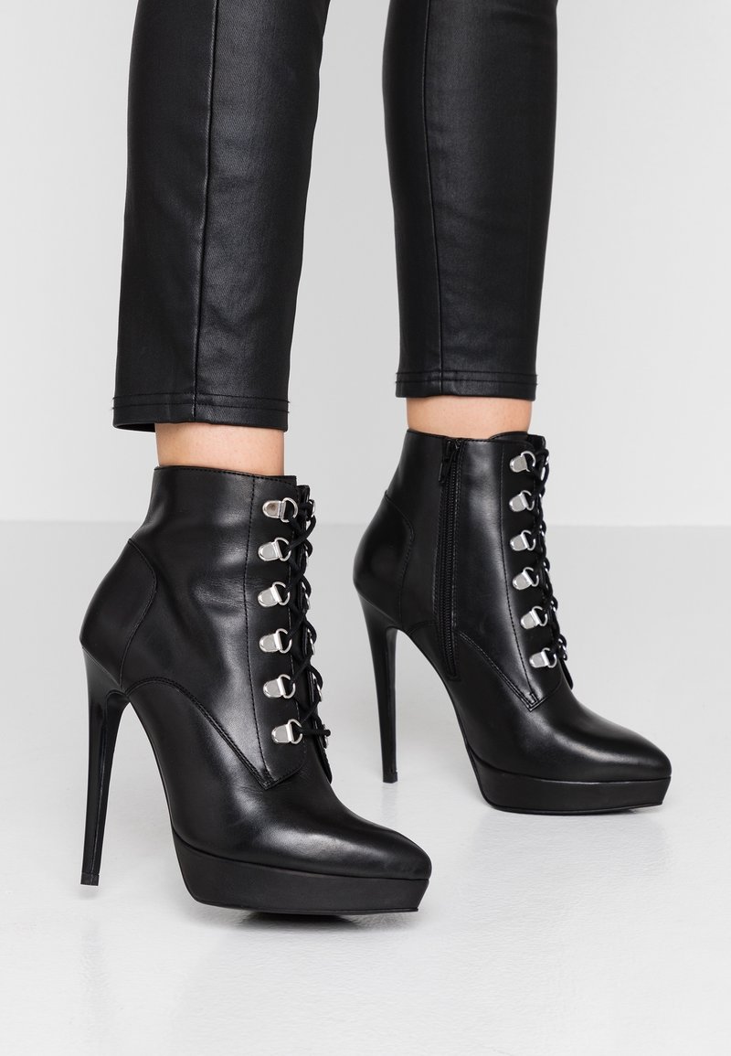Bottines noires à talons hauts avec lacets à l'avant et fermeture éclair sur le côté, portées avec un pantalon en cuir noir ajusté coupé au-dessus des chevilles.