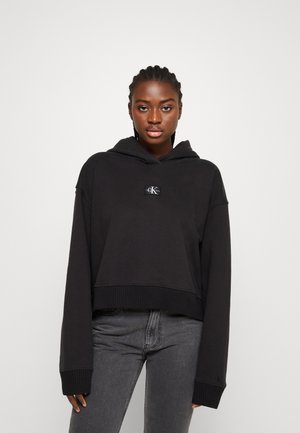 Sort cropped hoodie med et logo patch på forsiden, brede ærmer samt ribkant ved bund og manchetter. Fremstillet af blødt stof med en glat struktur.