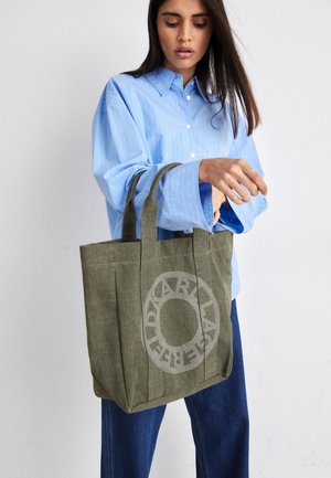 Woman in blue shirt and jeans holding an olive green tote bag with circular light print design, standing against a plain white background.