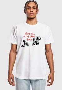 Hvid bomulds T-shirt med teksten "VI ER ALLE I DEN SAMME BÅD" i rød, ledsaget af sorte grafiske illustrationer af figurer.