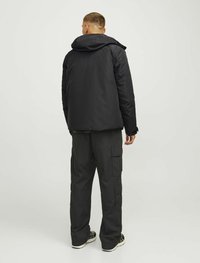 Chaqueta negra con capucha vista desde atrás, con un ajuste relajado. Llevada con pantalones cargo a juego, que muestran múltiples bolsillos laterales.