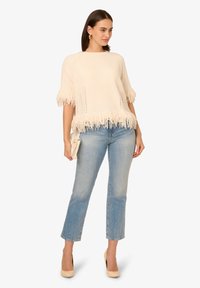 Cremfarbener gestrickter Oberteil mit Fransen an den Ärmeln und am unteren Saum, kombiniert mit hellblauen geraden Jeans und beigen High Heels.