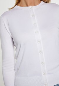 Witte cardigan gemaakt van zachte stof, met een ronde halslijn, lange mouwen en een rij van zes witte knopen aan de voorkant.