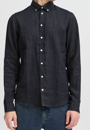 Homme portant une chemise noire à manches longues avec des boutons blancs et un jean bleu, debout devant un fond uni.