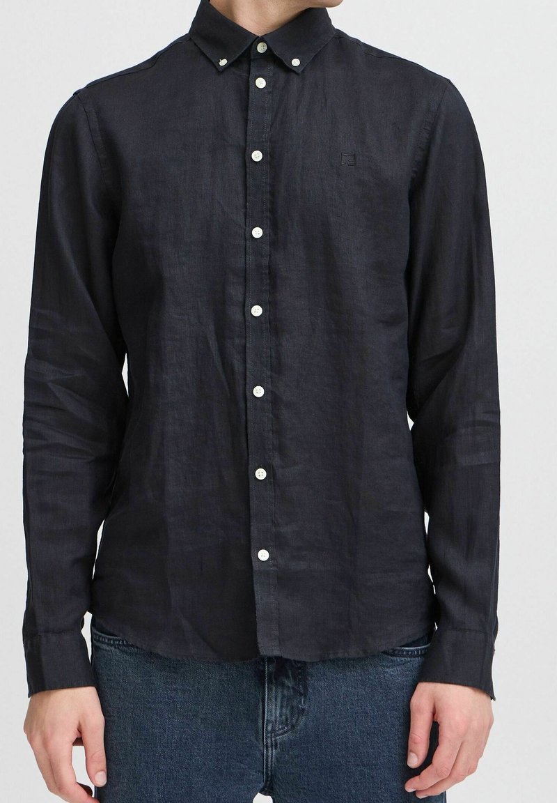 Homme portant une chemise noire à manches longues avec des boutons blancs et un jean bleu, debout devant un fond uni.
