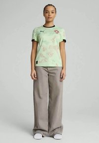 Zaļš īsām piedurknēm krekls ar teksturētu rakstu un melniem akcentiem, ar logotipu uz krūtīm. Kombinēts ar brīvām pelēkām biksēm un baltām kedām.