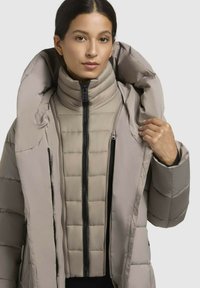 Doudoune beige clair, avec un col haut, un design piqué et des fermetures éclair doubles. L'intérieur présente une texture matelassée et un ajustement confortable.