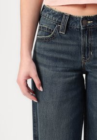 Temno pranje jeans z ravnim krojem, ki imajo visok pas, subtilne šive in konture žepov. Roka položena na tkanino.
