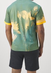 Grön mönstrad t-shirt med gula accenter, prydd med ett blommönster. Tyget har en vertikal randdesign, korta ärmar och en rundad fåll.