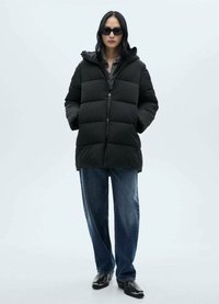 Zwarte pufferjas met horizontale gestikte secties, ritsafsluiting en capuchon. Gecombineerd met losse blauwe jeans en zwarte enkellaarsjes.