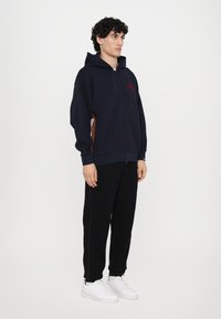 Hooded zip-up sweatshirt i marinblått med bruna sidopaneler, har elastiska mudd och nederkant. Kombinerad med svarta sweatpants och vita sneakers.