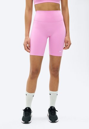 Kobieta ubrana w różowe, wysokie spodenki rowerowe, dopasowany krótki top, białe skarpetki Nike oraz czarne buty sportowe Nike, stoi na białym tle.