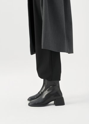Bottines en cuir noir à talons blocs bas portées avec un pantalon noir et un long manteau gris foncé.