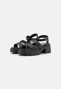 Sandales à plateforme noires avec de larges brides et une boucle de cheville sécurisée. Semelle épaisse avec un fini texturé et un design de crampons proéminents.