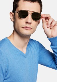 Okrągłe okulary przeciwsłoneczne z ciemnymi soczewkami i cienką, złotą metalową oprawką, trzymane przez osobę w jasnoniebieskim swetrze z dekoltem w serek.