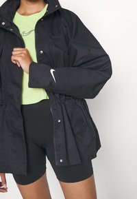 Nike svart vindjacka med dragkedja fram, elastisk midja och logotyp på ärmen, i kombination med en grön topp och svarta shorts.