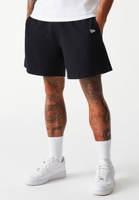 Pantaloni corti neri in felpa con tasche laterali, vita elastica e un piccolo logo ricamato. Indossati con sneakers bianche e calze bianche a crew.