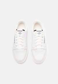 Tênis brancos de couro com uma textura suave, apresentando ponta perfurada, cadarços planos e a marca Reebok na lingueta e na lateral.