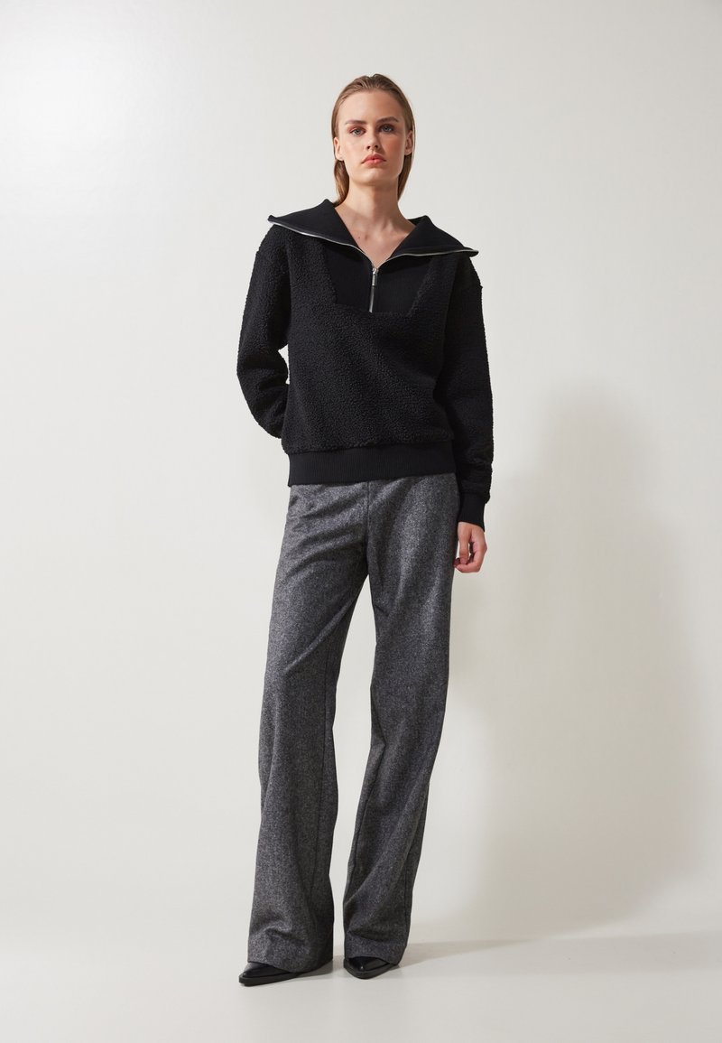 Pullovre en polaire noir avec une fermeture éclair quart et un large col, associé à un pantalon large gris avec un motif texturé.