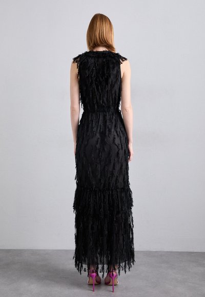 Robe noire avec des détails à franges, corsage ajusté et jupe à volants. Portée avec des chaussures à talons roses, vue de dos sur un fond uni.