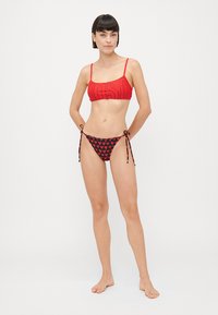 Sarkans bikini tops ar izteiksmīgu logotipu, savienots ar melniem bikini apakšiem ar sarkano sirds rakstu un regulējamiem sānu saitēm.