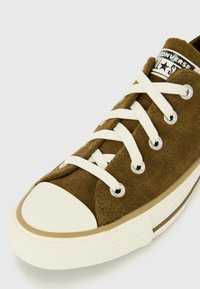 Zapatillas de ante marrón con cordones blancos y puntera de goma. Presenta una suela de goma texturizada y una etiqueta de logo en la lengüeta.