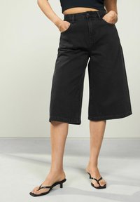 Černé denimové culottes s vysokým pasem, širokými nohavicemi a předními kapsami. Stylizováno s černými sandály na podpatku na světle šedé podlaze.