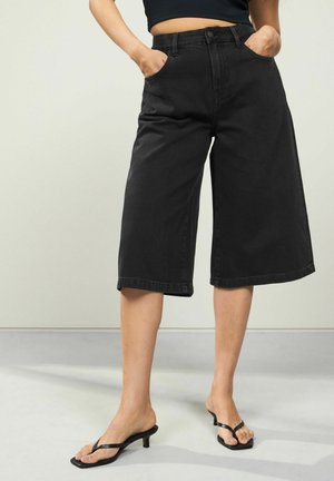 Schwarze Denim-Culottes mit hoher Taille, weitem Bein und Fronttaschen. Gestylt mit schwarzen, hochhackigen Flip-Flops auf einem hellgrauen Boden.