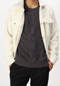 Biała kurtka z polaru z srebrnymi zatrzaskami, noszona na ciemnoszarym dzianym swetrze z fakturowanym wzorem i ściągaczem na dole.