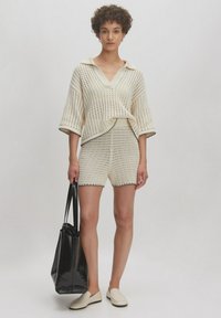 Beige gebreide top met een V-hals en korte mouwen, gecombineerd met bijpassende shorts. Zwarte accenten en een zwarte tote bag maken de look compleet.
