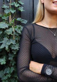 Črna prozorna bluzka z dolgimi rokavi in črnimi pikami, oprijete oblike, kombinirana s srebrno uro z okroglim zaslonom in mrežastim paščkom. V ozadju zelena bršljanova rastlina.