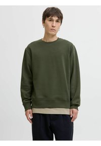 Olijfgroene sweatshirt van zacht materiaal met een ronde hals, geribbelde manchetten en zoom, gelaagd over een beige T-shirt, gedragen met donkere broek.