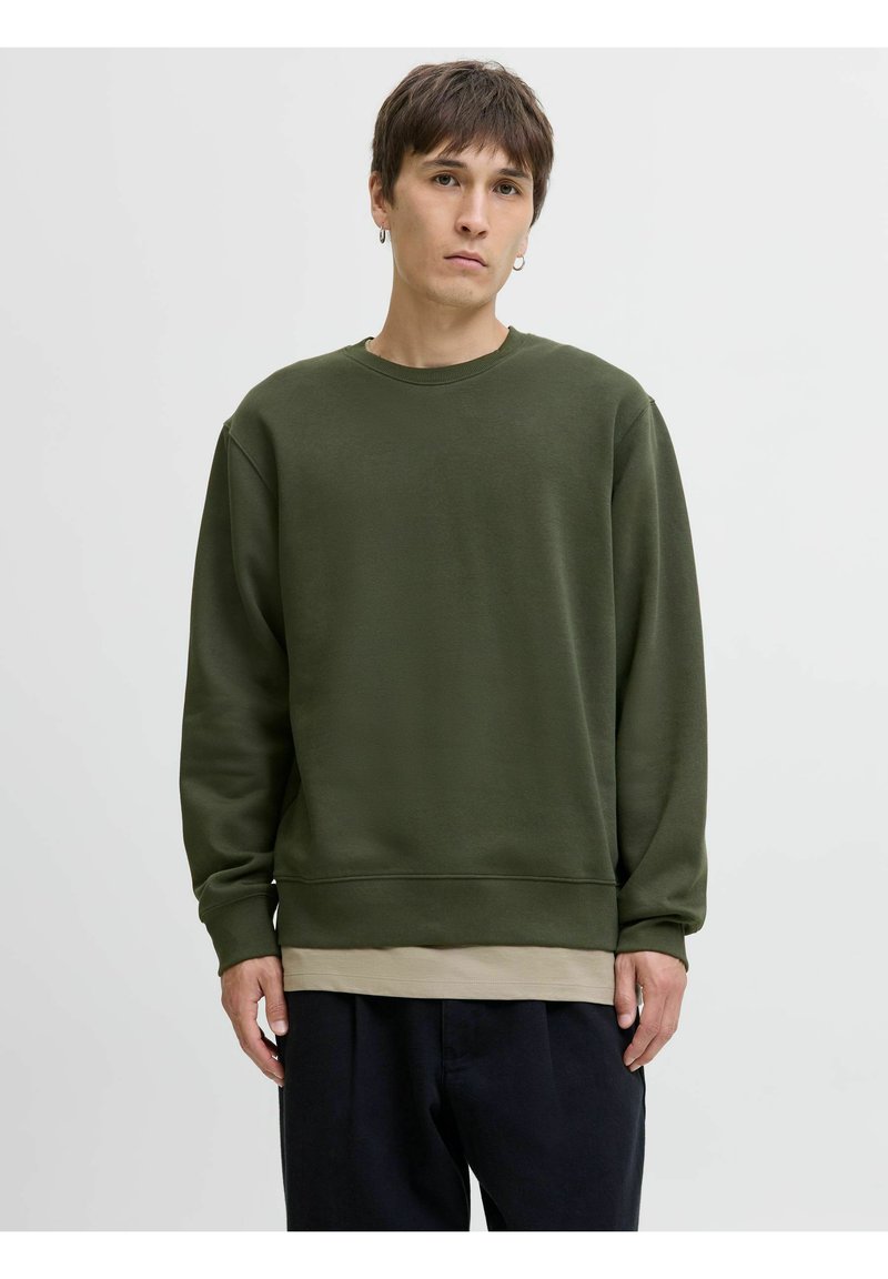 Sudadera verde oliva de tela suave con cuello redondo, puños y dobladillo acanalados, superpuesta sobre una camiseta beige, combinada con pantalones oscuros.