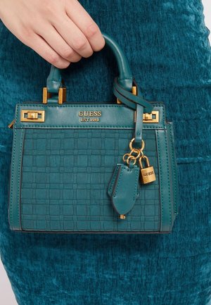 Hand die een teal Guess-handtas vasthoudt met geweven structuur, gouden beslag en hangend slotje als hanger tegen een bijpassende teal stoffen achtergrond.