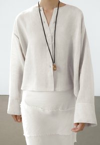 Blouse ample beige clair avec patte de boutonnage, manches longues et texture douce, associée à une jupe blanche superposée.
