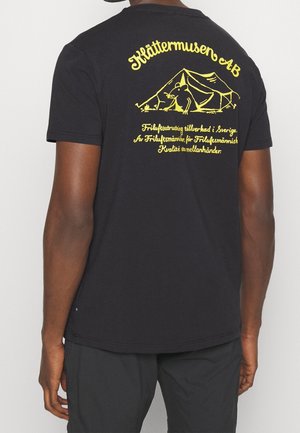 T-shirt en coton noir avec un imprimé graphique jaune représentant une tente et un animal au dos, avec le texte de la marque au-dessus et en dessous du design.