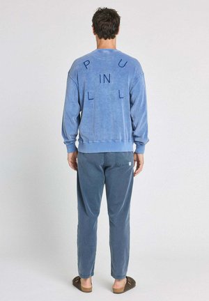 Un homme se tient dos à nous, portant un sweat-shirt bleu avec des lettres éparpillées "P U L L IN L" au dos, un pantalon bleu et des sandales marron.